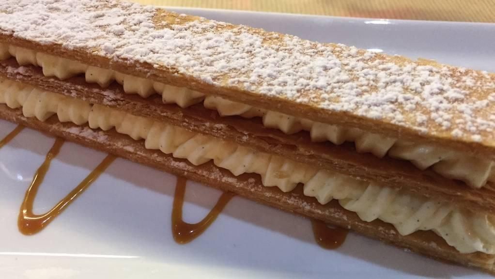 Pâtisserie mille-feuille à la garniture crémeuse, saupoudrée de sucre glace et nappée de caramel, présentée sur une assiette blanche.