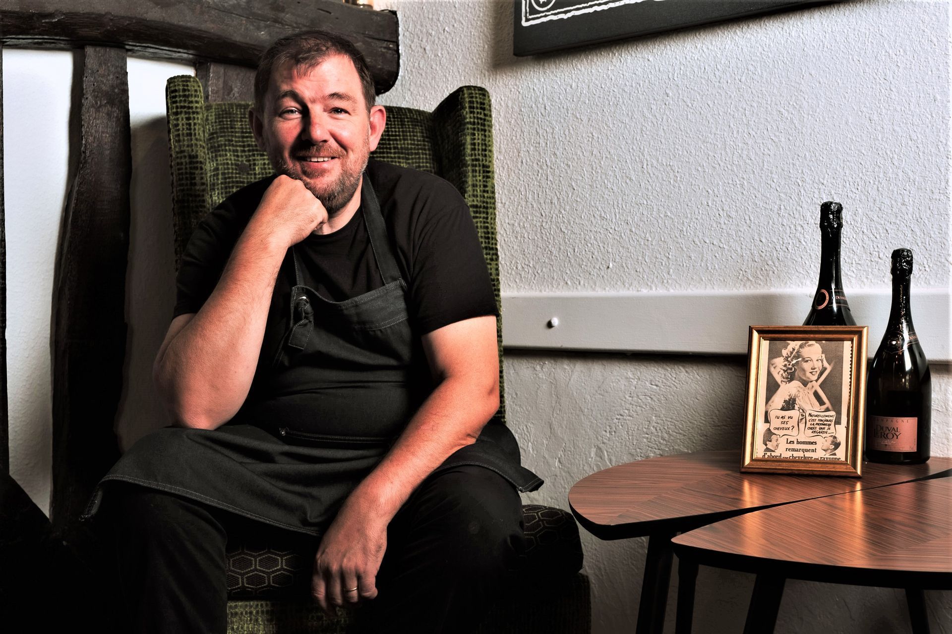 Un homme est assis sur une chaise, le menton appuyé sur sa main. Près d'une table avec des bouteilles, un tableau encadré et un mur texturé.