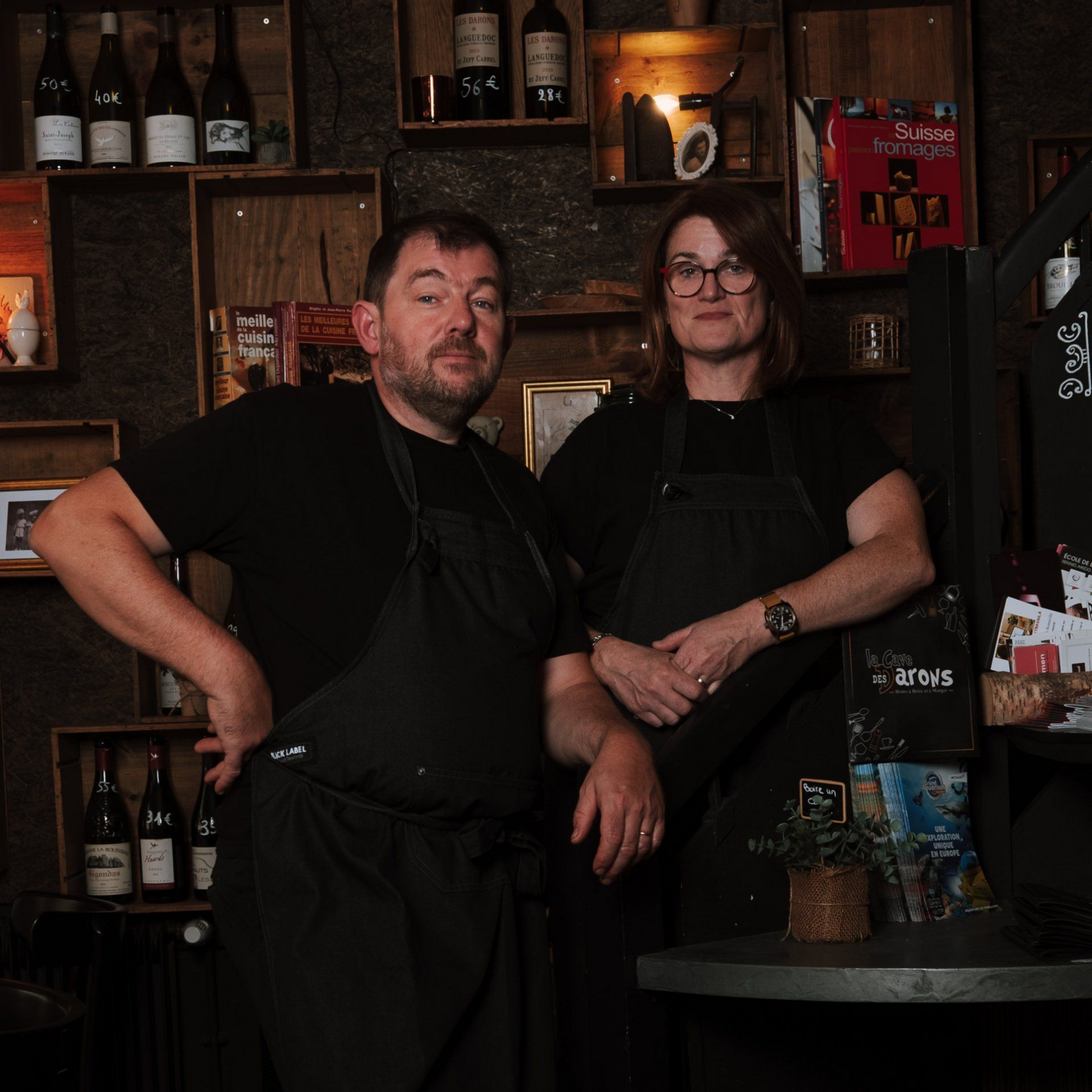 Deux personnes, vêtues de tabliers noirs, se tiennent à l'intérieur, près d'une table. Des étagères remplies de bouteilles sont visibles en arrière-plan.
