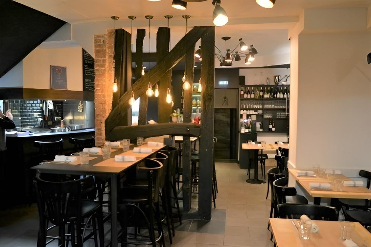 Intérieur de restaurant avec tables hautes, chaises noires, poutres apparentes et éclairage d'ambiance.