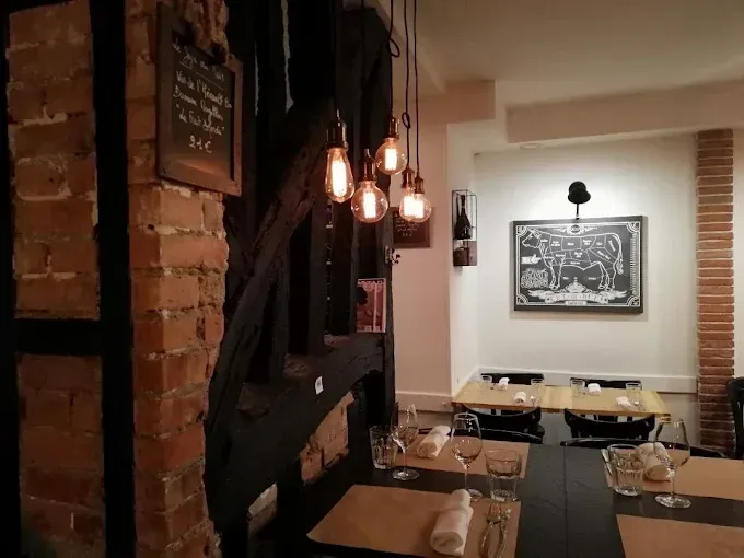 Intérieur du restaurant avec briques apparentes et poutres en bois sombre. Tables dressées et éclairage par ampoules Edison.