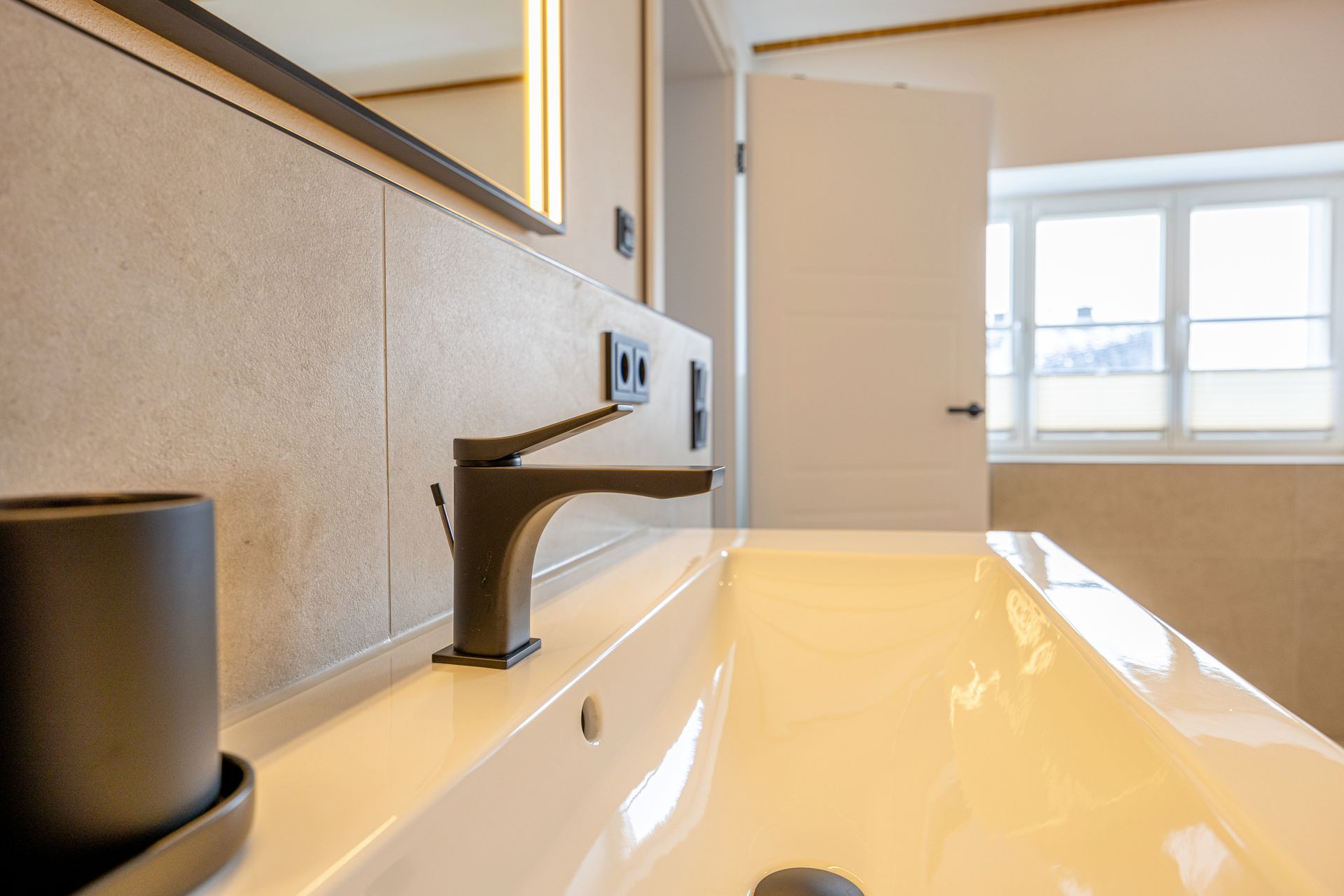 Waschbecken mit schwarzem Wasserhahn, schwarzem Zahnputzbecher und weißem Porzellanbecken. Die Tür ist offen.