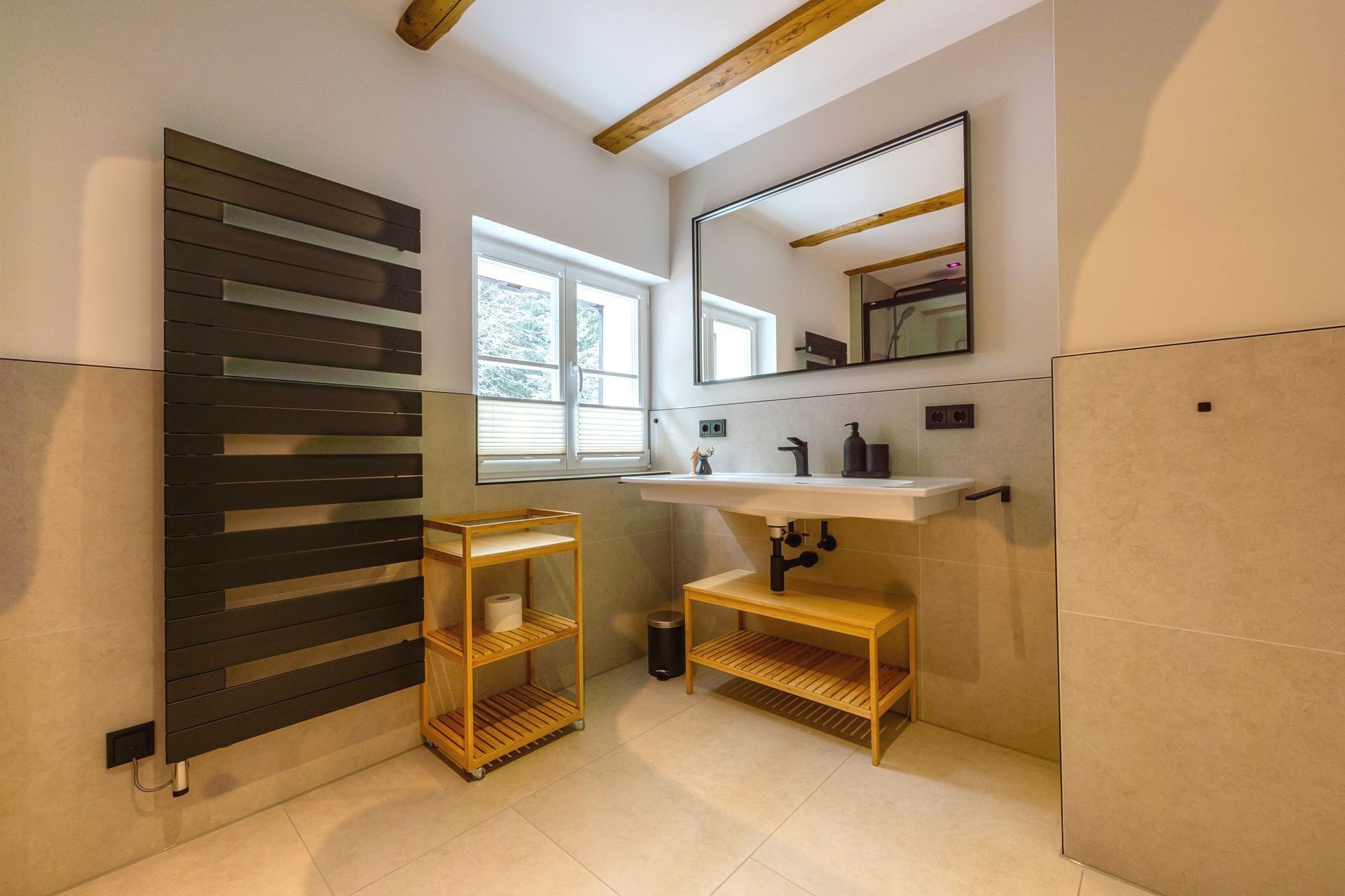 Badezimmer mit großem Spiegel, weißem Waschbecken, Holzregalen und dunklem Heizkörper. Helle Wände und Boden.