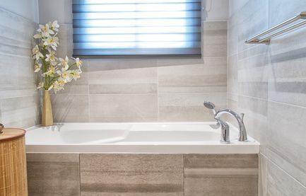 Modernes Badezimmer mit weißer Badewanne, hellgrauen Fliesen und einem kleinen Fenster mit Jalousien.