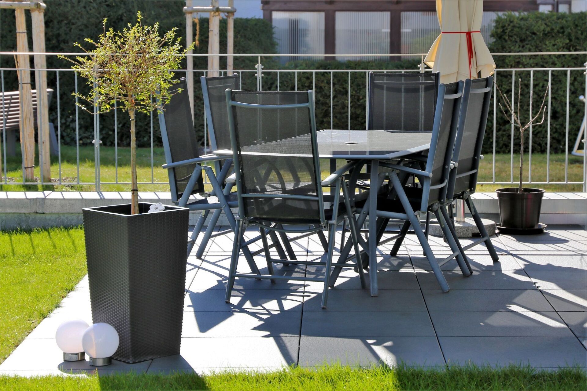 Terrasse mit Tisch, Stühlen und einem kleinen Baum in einem hohen schwarzen Topf. Sonniger Tag.