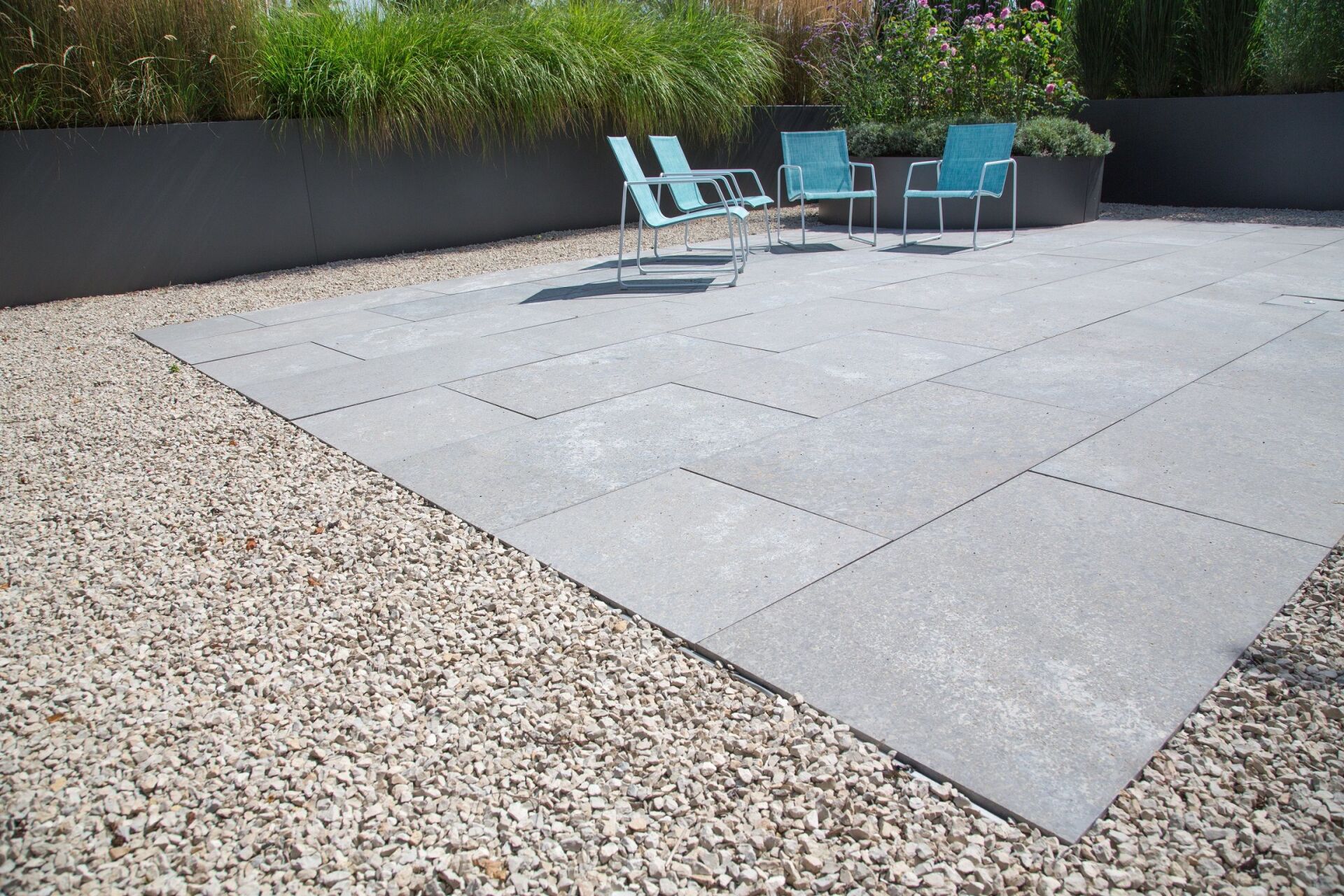 Steinterrasse mit hellblauen Stühlen, umgeben von Kies und Pflanzen.