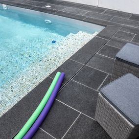 Poolrand mit dunklen Fliesen, Stufen und zwei Poolnudeln (grün und lila) sowie einem grauen Kissen.