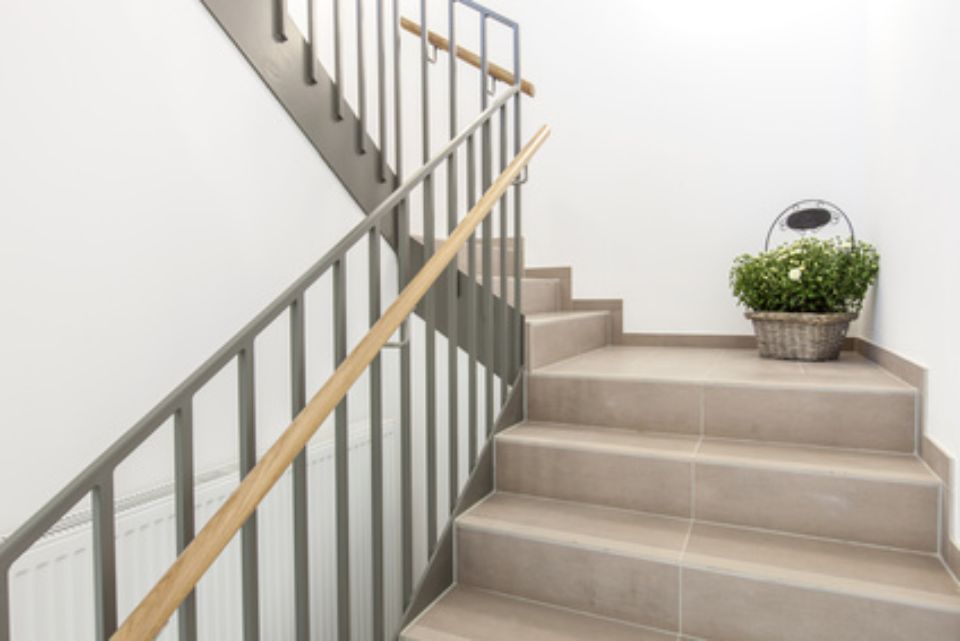Treppe mit hellgrauen Stufen, Metallgeländer, Holzhandlauf und einem Korb mit Pflanzen vor einer weißen Wand.