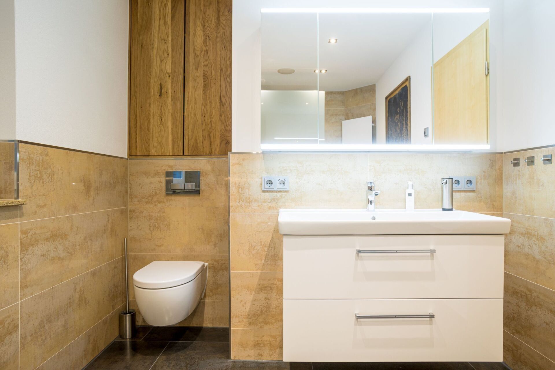 Modernes Badezimmer mit weißem Waschtisch, Toilette und Fliesen in warmen Farbtönen. Holzschrank über der Toilette.