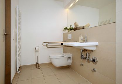 Barrierefreies Badezimmer mit wandmontierter Toilette und Waschbecken sowie Haltegriffen für mehr Mobilität. Beige Fliesen und Wände.