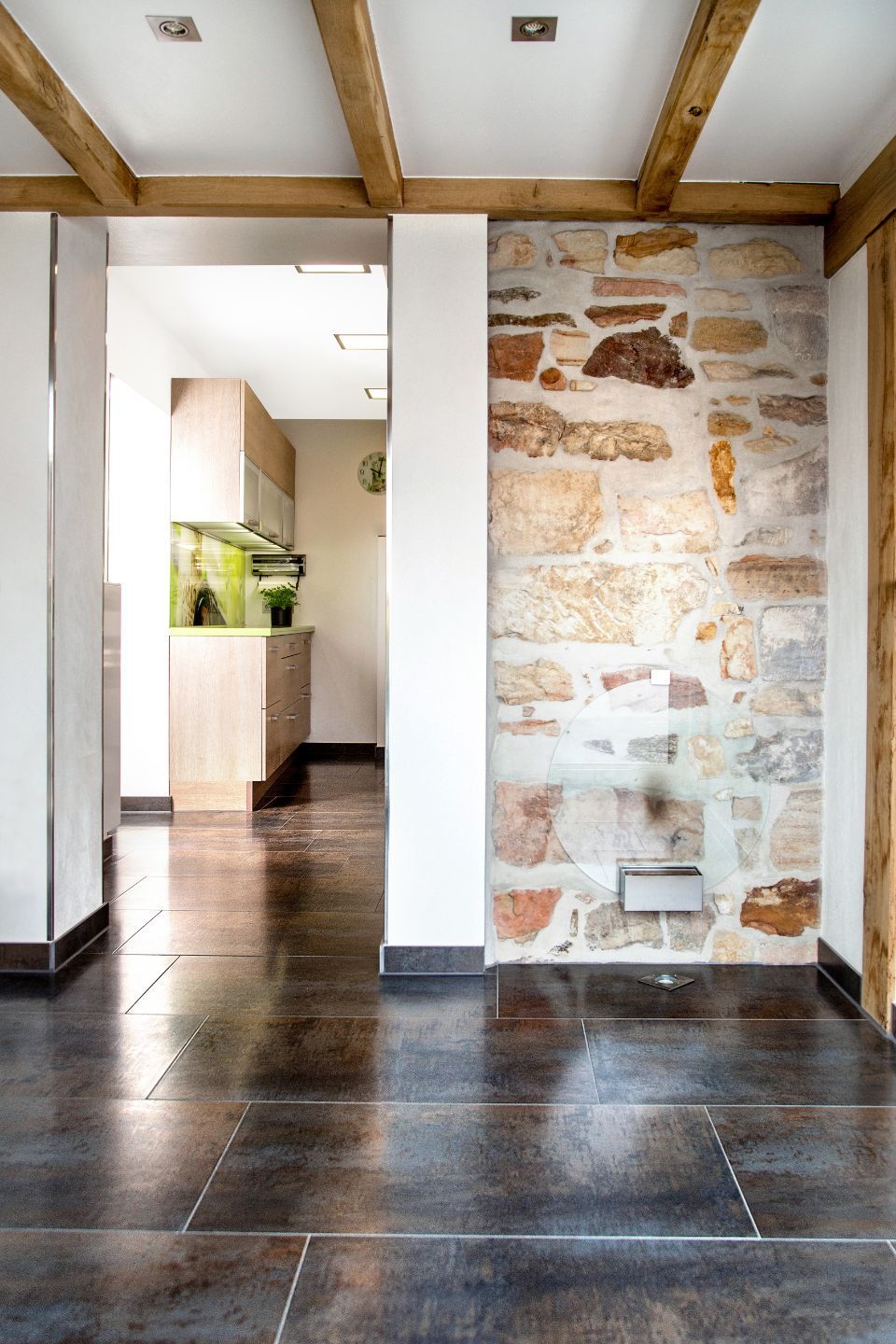 Steinwand in einem Raum mit glänzendem dunklem Boden, der zu einer Küche führt. Holzbalken und weiße Säulen.