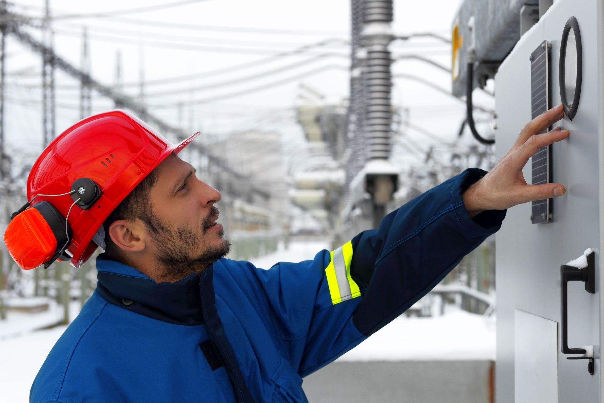 Trabajador electricista con casco rojo, chaqueta azul y protección auditiva, revisando equipos en una central eléctrica.