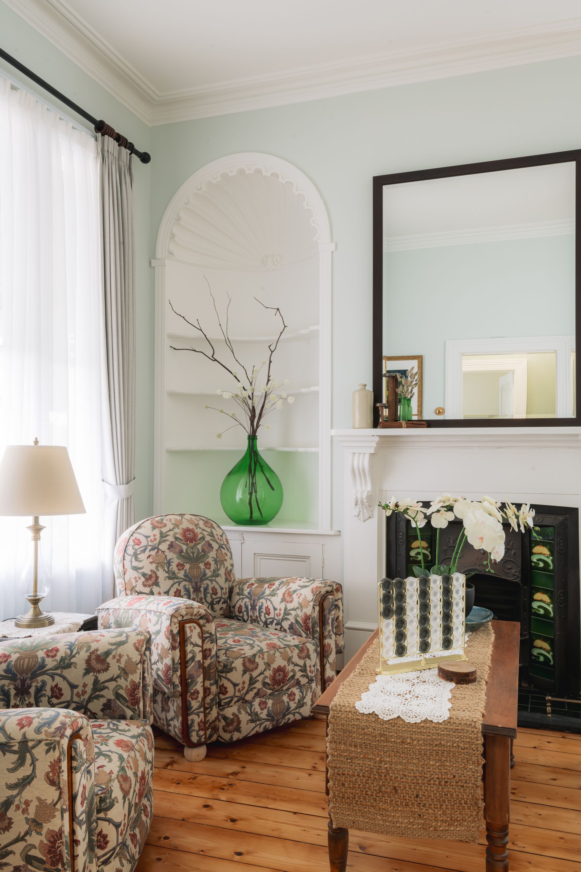 A living room with two chairs and a fireplace.  Green vase, Floral couch, Victorian detail.