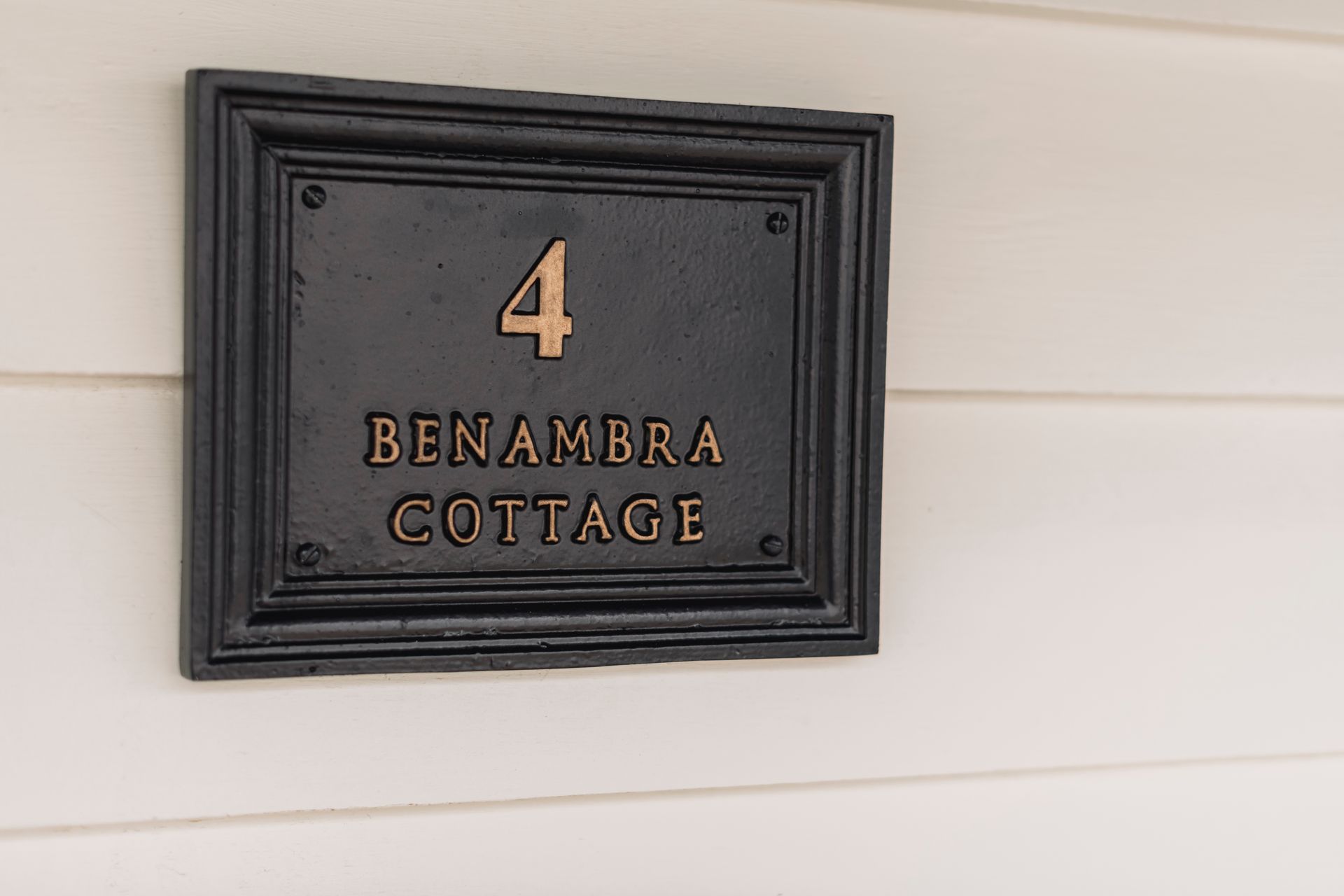 A black and gold sign on a white wall says benambra cottage.