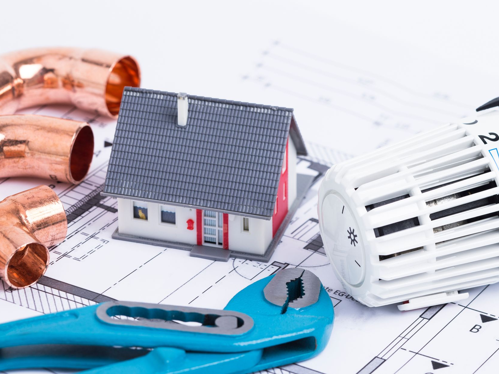 Maquette de maison posée sur des plans techniques, entourée d’un robinet thermostatique, d’un outil de plomberie bleu et de tubes en cuivre
