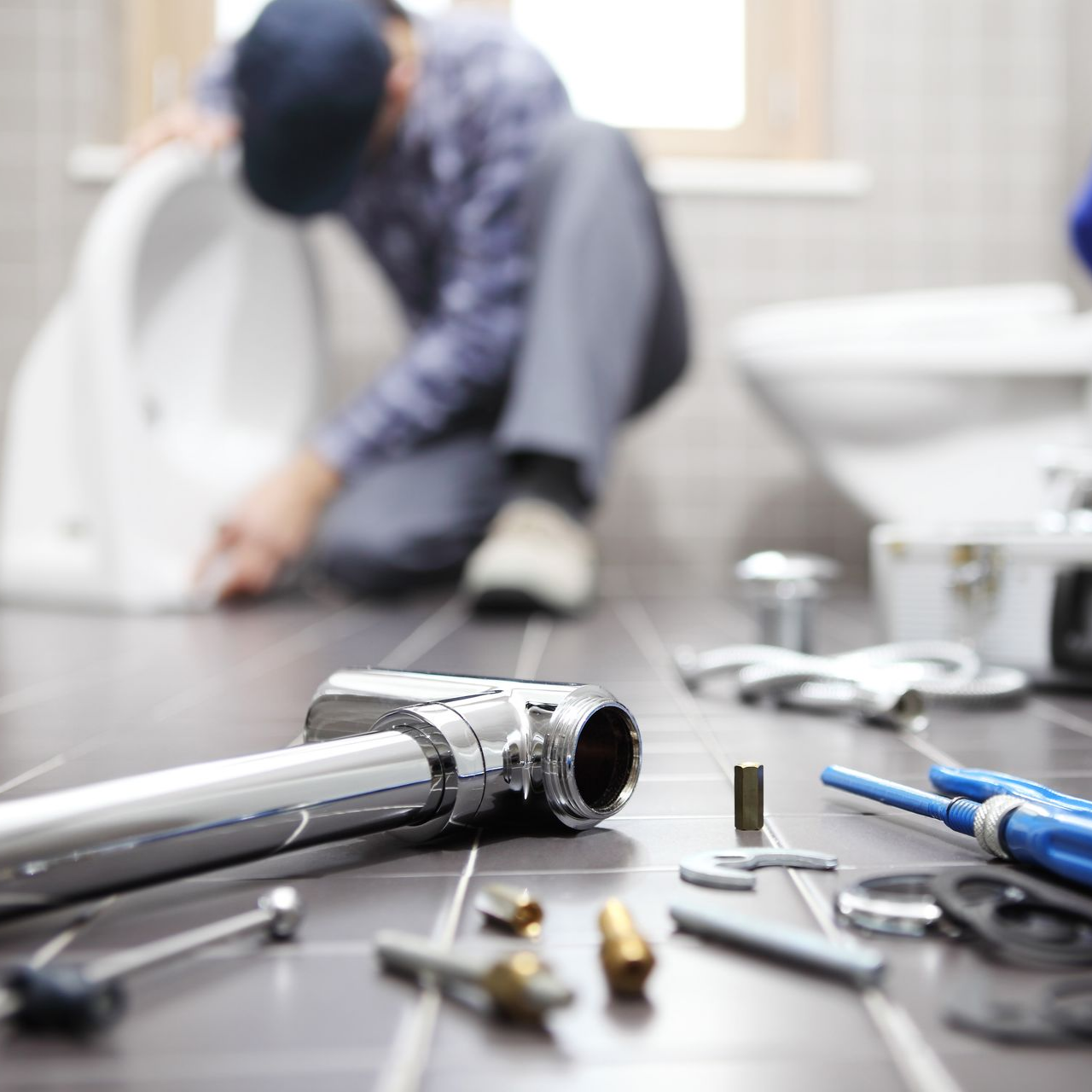 Gros plan sur un tuyau chromé et des outils de plomberie posés au sol, avec un plombier travaillant en arrière-plan dans une salle de bains