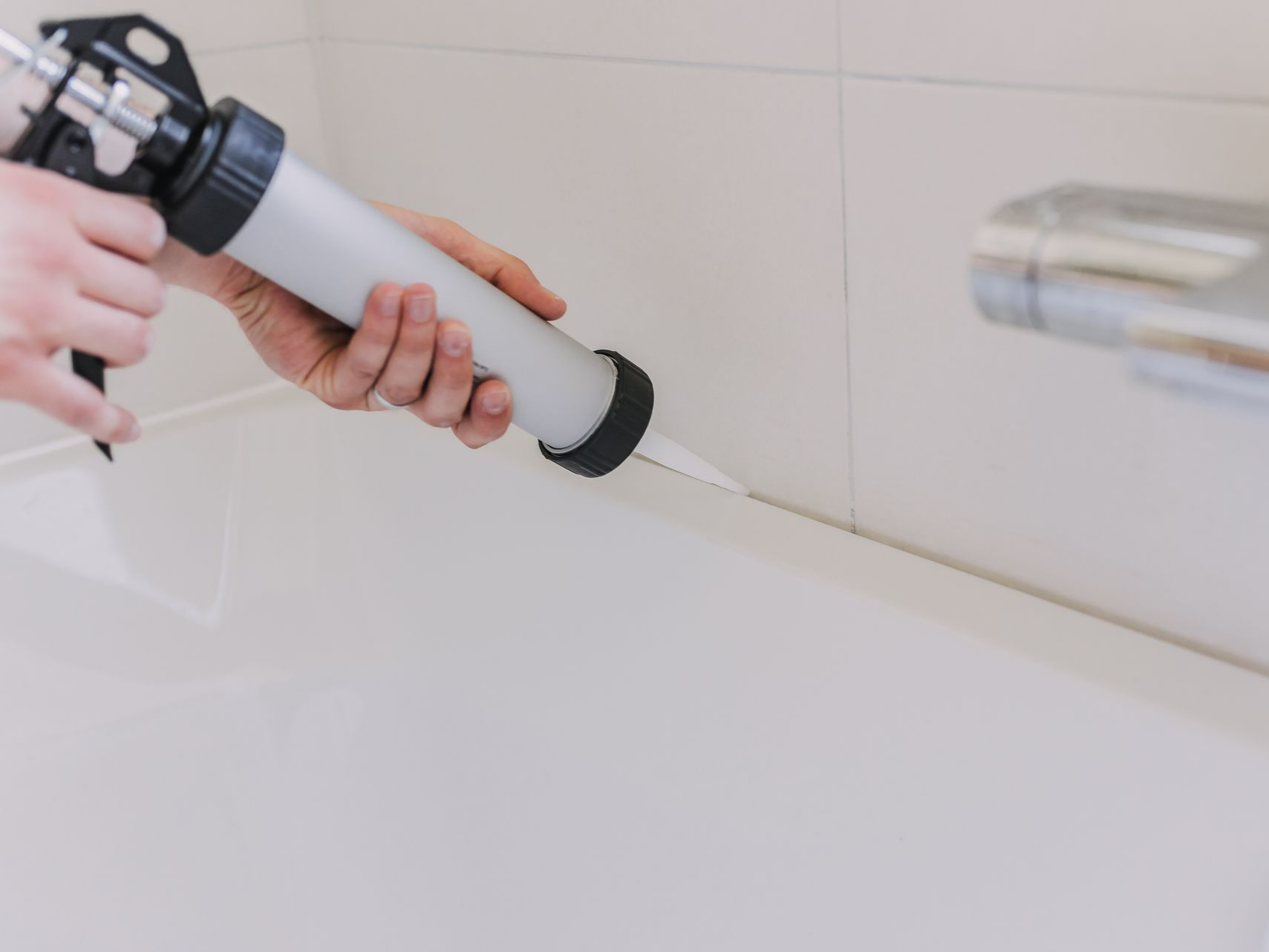 Main appliquant un joint de silicone avec un pistolet sur le bord d’une baignoire