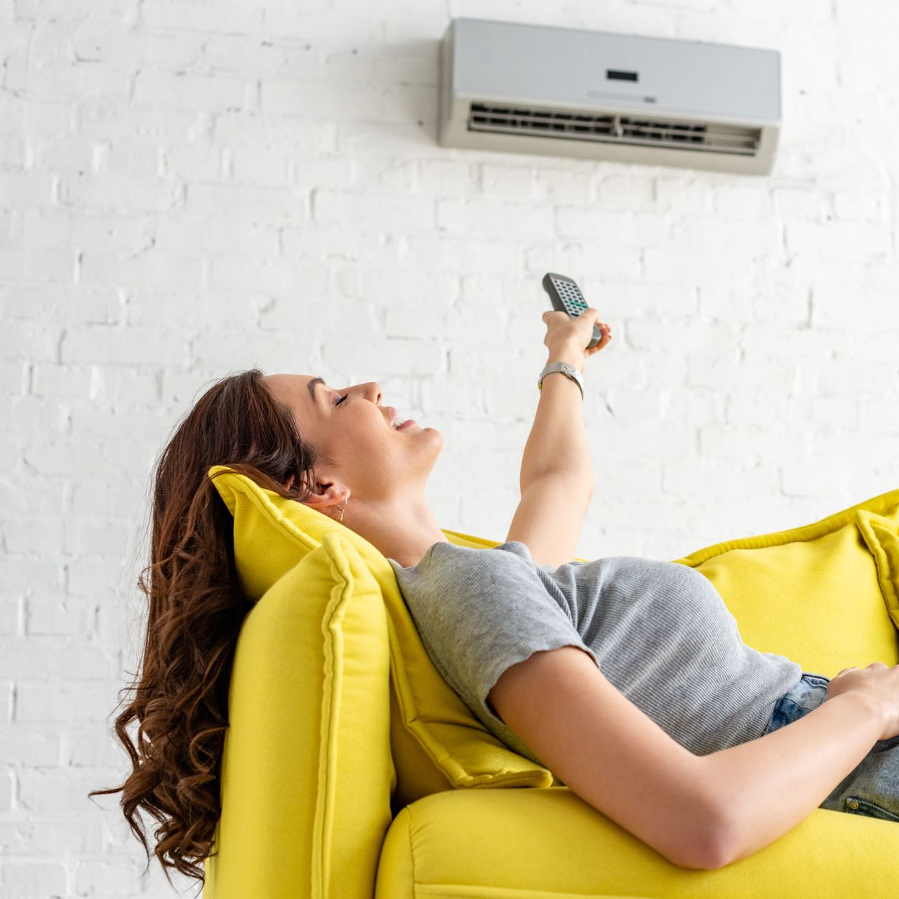 Personne allongée sur un canapé jaune utilisant une télécommande pour régler un climatiseur mural