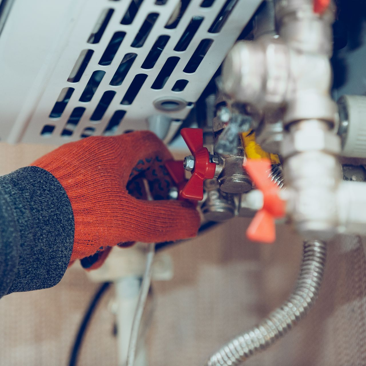 Main portant un gant rouge ajustant une vanne sur une chaudière avec plusieurs tuyaux et raccords visibles