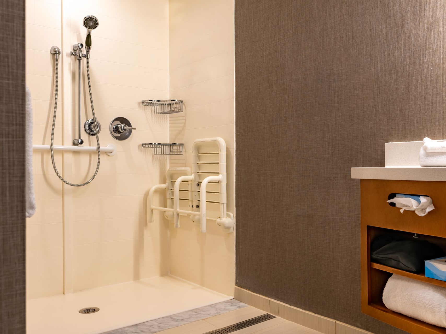 Douche à l’italienne équipée de barres d’appui murales, siège rabattable et étagères métalliques pour salle de bain accessible