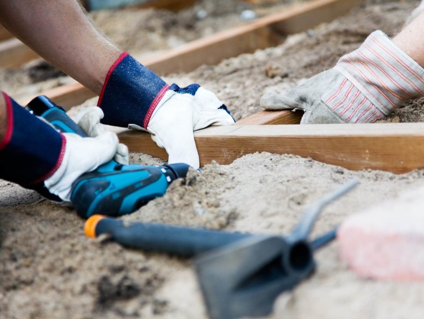 Hände mit Handschuhen benutzen einen Bohrer, um Holzbalken auf einem sandigen Untergrund zu befestigen.