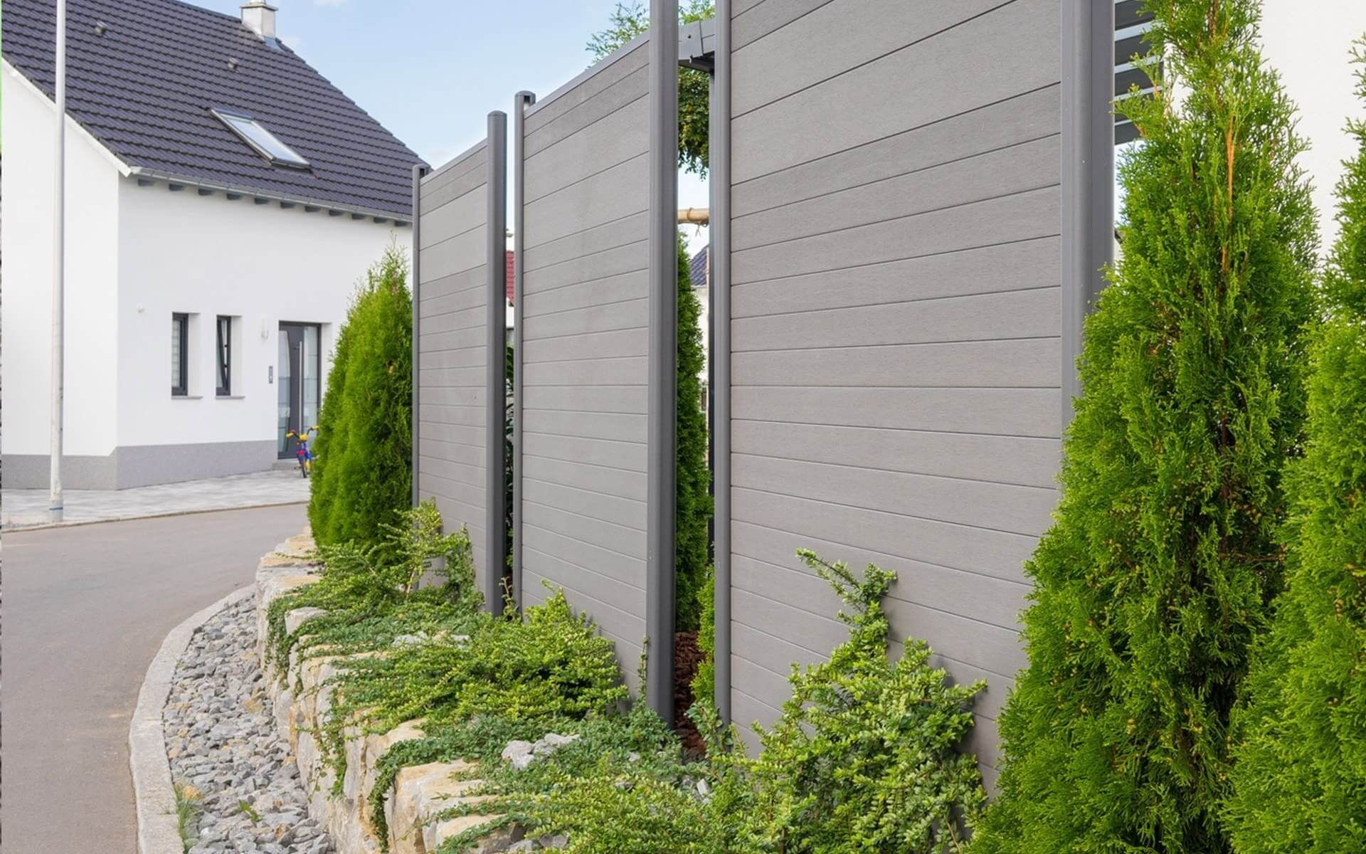 Grauer Sichtschutzzaun mit horizontalen Linien, neben einer Steinmauer und Grünpflanzen.