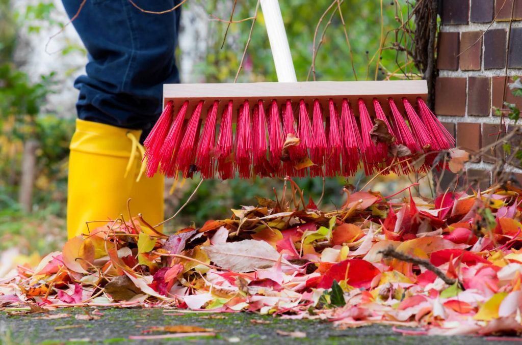 Eine Person in gelben Stiefeln kehrt mit einem roten Besen bunte Herbstblätter zusammen.