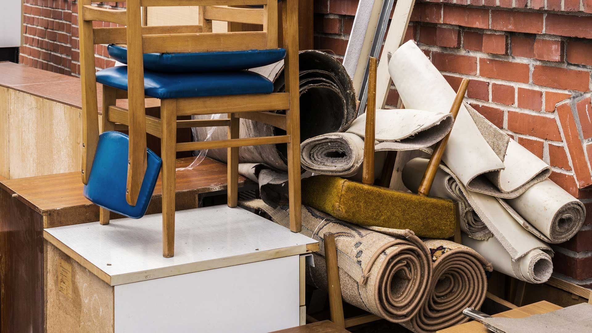 Ein Stapel zusammengerollter Teppiche, Möbel und Reinigungsmittel lehnt an einer Backsteinmauer.