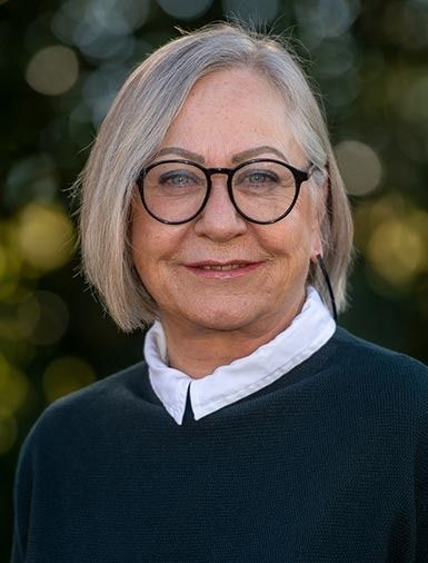 Frau mit grauem Bob-Haarschnitt, Brille und einem dunkelgrünen Pullover über einem weissen Hemd mit Kragen, lächelt im Freien.