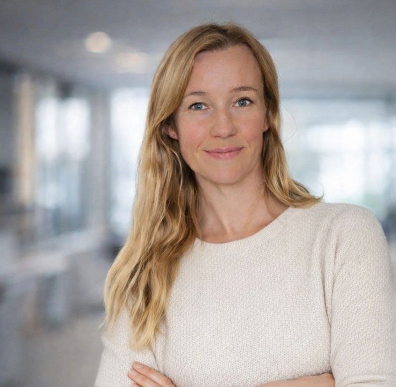 Eine Person mit langen blonden Haaren, die einen beigen Pullover trägt und in einem Büro mit verschränkten Armen posiert.