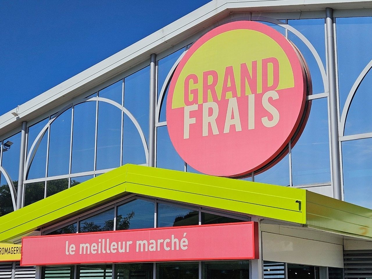 Grand Frais store with a large round sign, green and pink, under a blue sky.