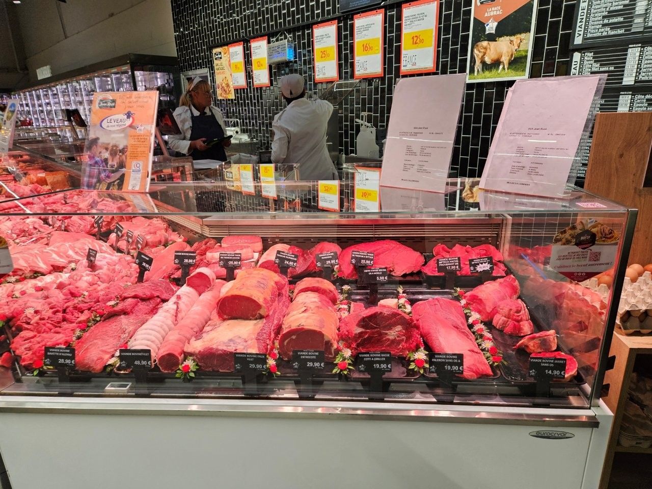 Eine Fleischereitheke mit verschiedenen Stücken rotem Fleisch, im Hintergrund zwei Mitarbeiter.