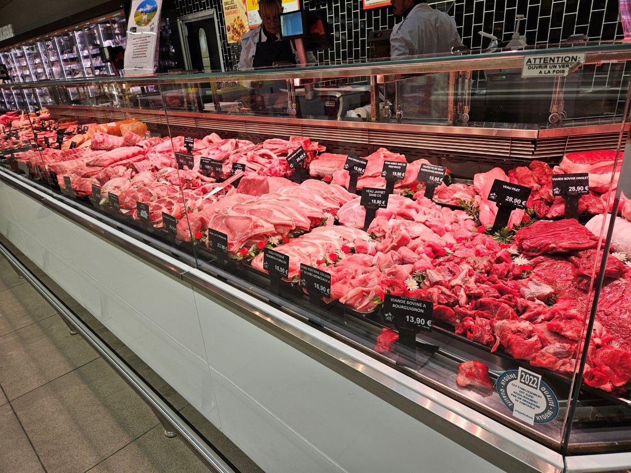 Eine Fleischereitheke mit verschiedenen Stücken rotem Fleisch, im Hintergrund zwei Mitarbeiter.