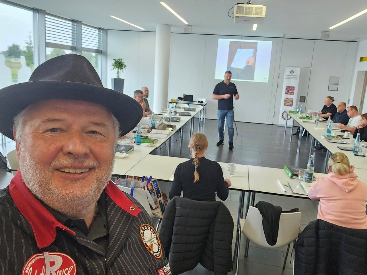 Mann mit Hut macht Selfie in einem Schulungsraum; Moderator vorne, Teilnehmer sitzen an Tischen.