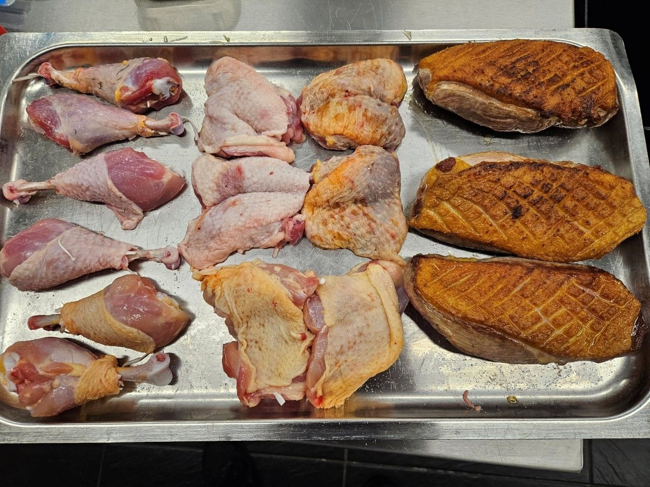 Auf einem Metalltablett liegen verschiedene rohe Ententeile: Keulen, Schenkel und Brust, gewürzt und kochfertig.