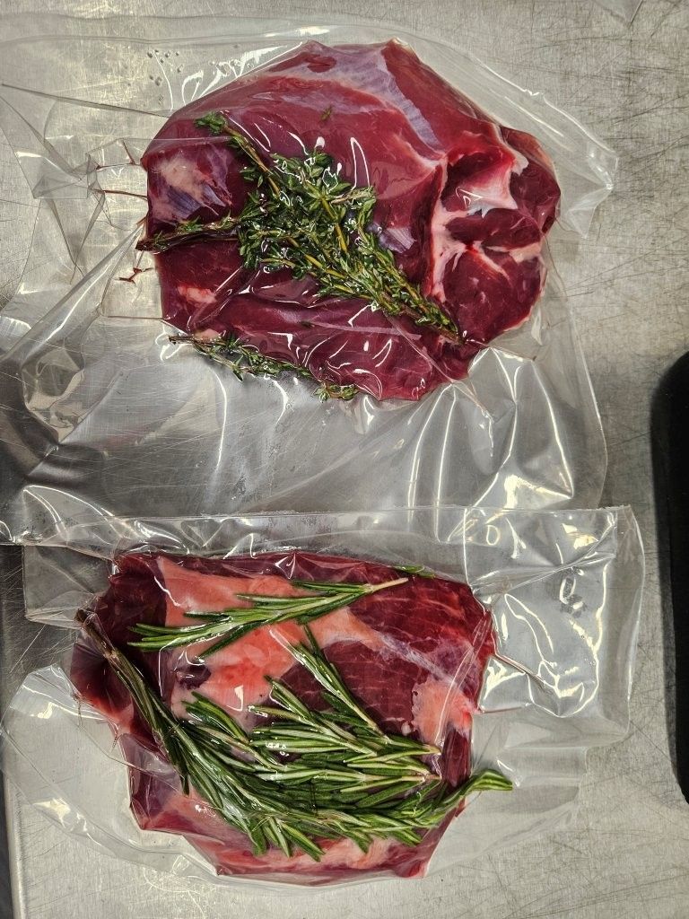 Zwei rohe, vakuumverpackte Fleischstücke, eines mit Thymian, eines mit Rosmarin, auf einer Metalloberfläche.