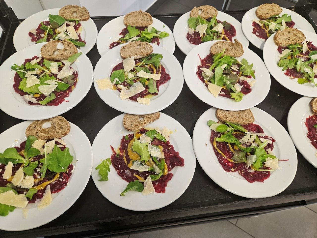Teller mit Rote-Bete-Carpaccio mit Rucola, Parmesan und einer Scheibe Brot, auf einem Tablett angerichtet.