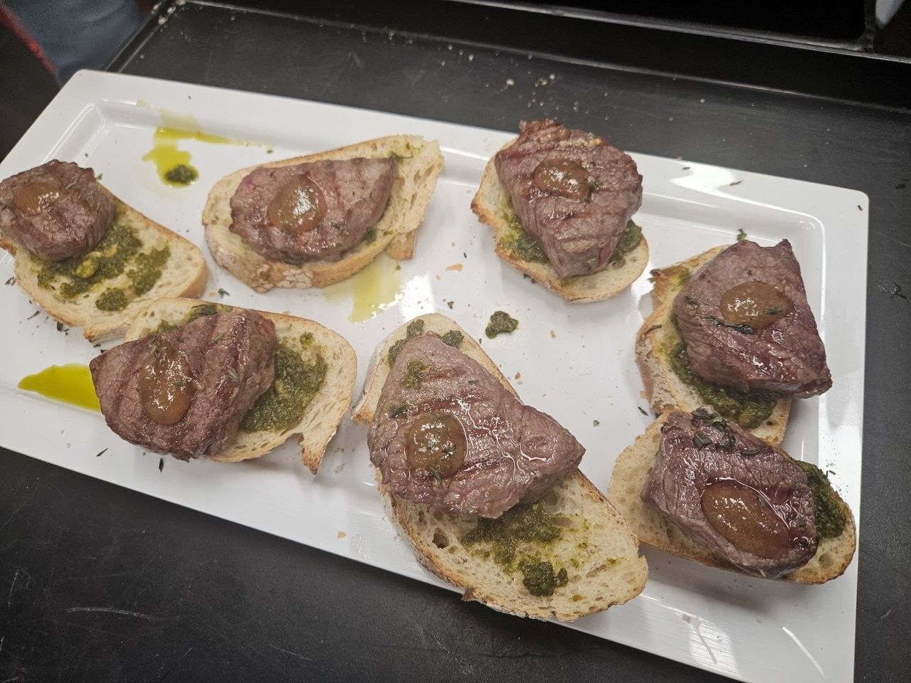 Bruschetta: Gegrillte Fleischscheiben auf geröstetem Brot mit Pesto, auf einem weißen rechteckigen Teller.