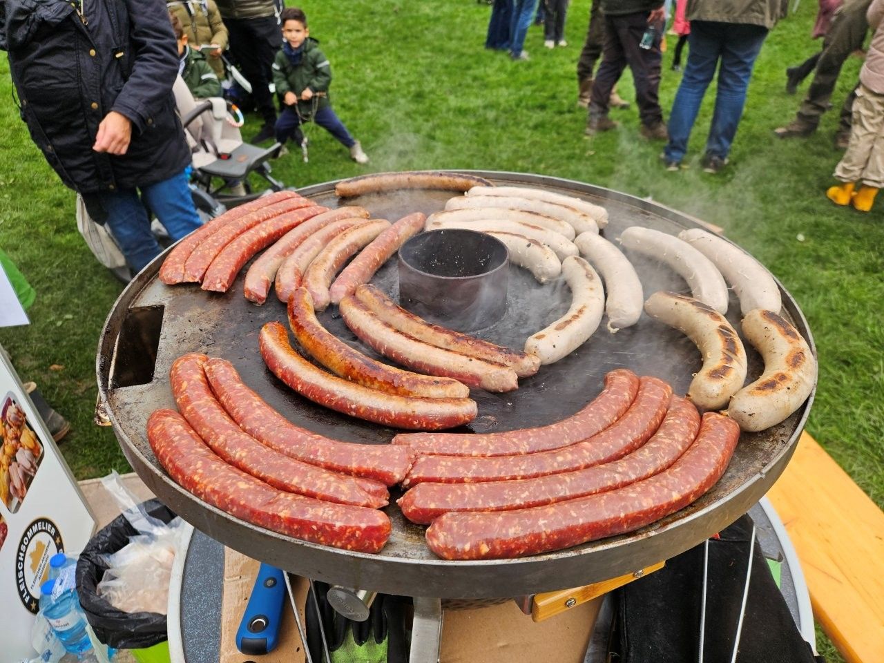 Gegrillte Würstchen auf einer großen runden Grillplatte bei einer Veranstaltung im Freien.