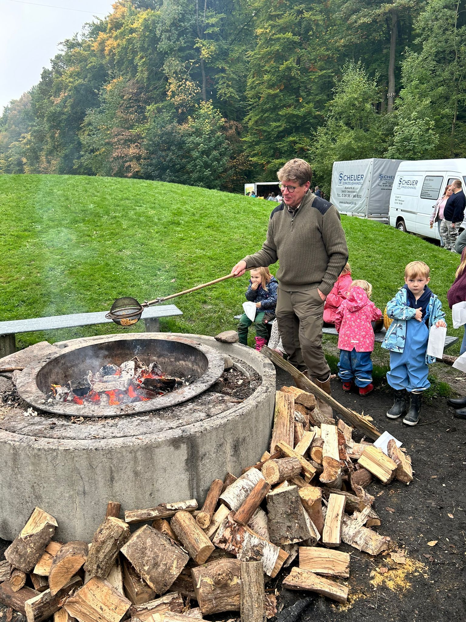 Mann rührt Topf über Feuer, Kinder schauen zu. Draußen, Brennholz, grünes Gras, Bäume im Hintergrund.
