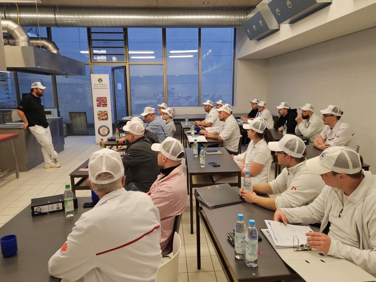 Ein Mann spricht in einer Küche zu einer Gruppe von Menschen mit weißen Kochmützen. Sie sitzen an Tischen und hören zu.
