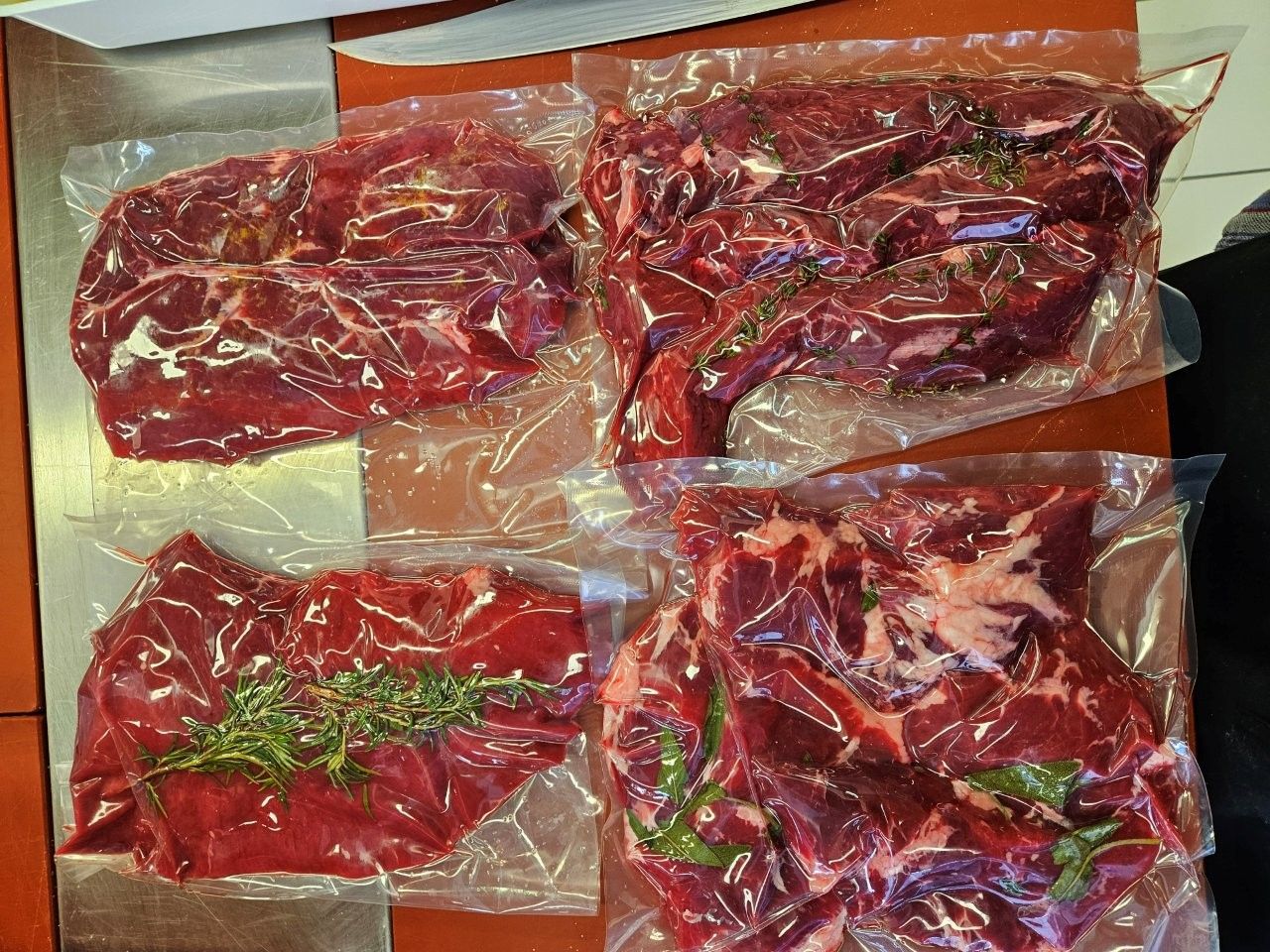 Vier vakuumverpackte Päckchen mit rohem rotem Fleisch, möglicherweise Steaks, auf einer roten Oberfläche mit Kräutern.