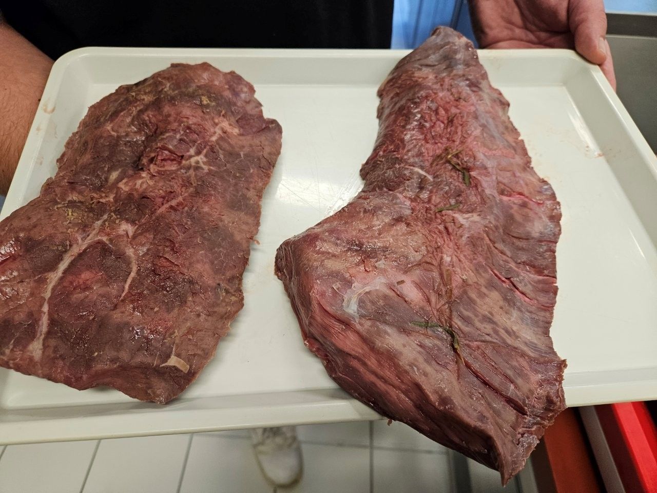 Zwei rohe Rindfleischstücke auf einem weißen Tablett, das von einer Person gehalten wird.
