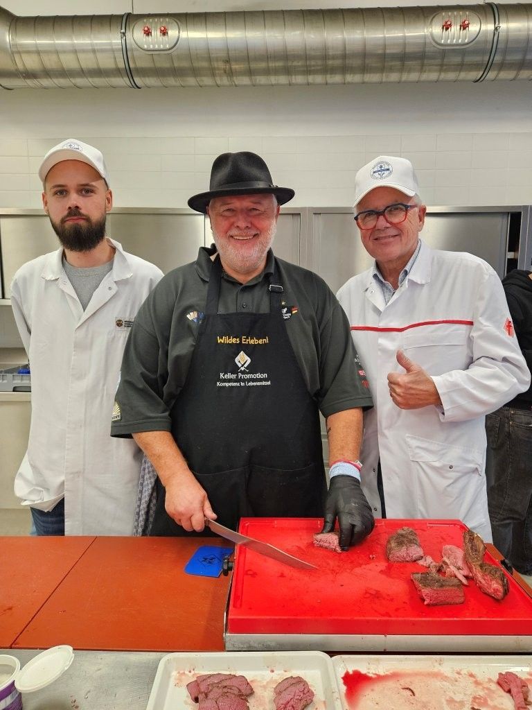 Drei Männer in der Metzgerei: einer schneidet Fleisch, einer zeigt den Daumen nach oben, einer beobachtet. Rotes Schneidebrett.