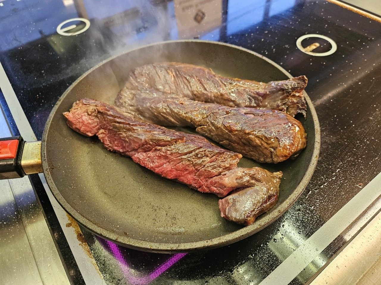 Zwei Steakstücke braten in einer Pfanne auf dem Herd, Dampf steigt auf.