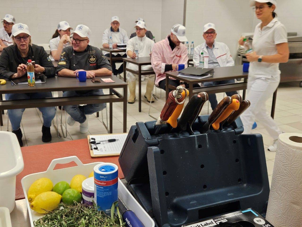 Kochkurs: Schüler in weißen Kochmützen und -mänteln hören dem Ausbilder zu. Messer und Zutaten liegen auf dem Tisch.