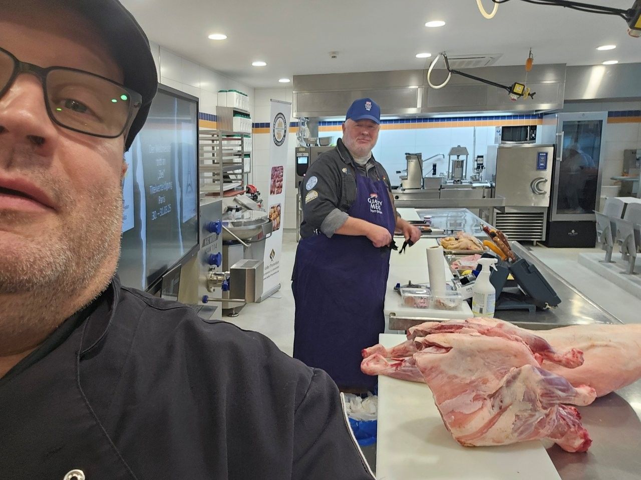 Zwei Metzger in einer Großküche, auf einem Tisch liegt rohes Fleisch. Einer macht ein Selfie, der andere arbeitet.