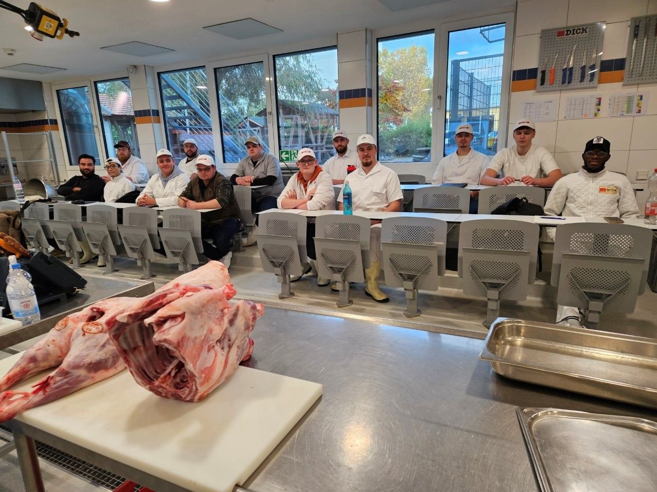 Fleischverarbeitungskurs: Schüler in weißen Kitteln und Hauben sitzen an Tischen. Rohes Fleisch liegt auf einem Tisch vor ihnen.
