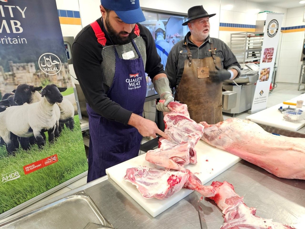 Zwei Metzger zerlegen Lammfleisch an einer Theke. Der eine trägt eine Schürze, der andere eine Mütze. Links hängt ein Banner.