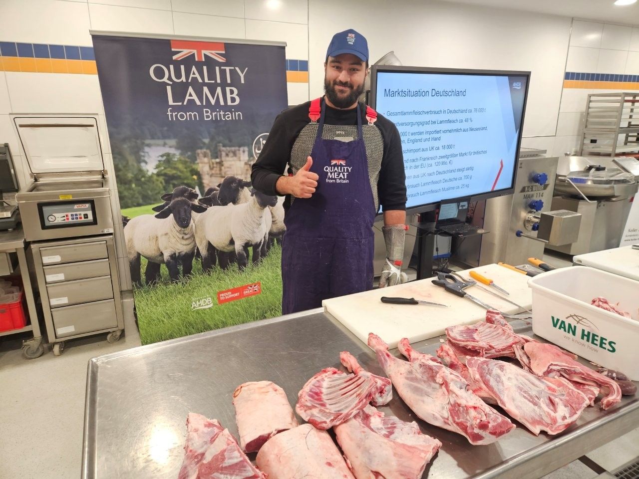 Ein Metzger demonstriert das Zerlegen eines Lamms. Er trägt Schürze und Mütze und zeigt den Daumen nach oben. Verschiedene Lammfleischstücke liegen auf einem Edelstahltisch.